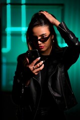 Stylish woman in black leather jacket using smartphone at night with neon light background