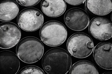 A monochrome close-up shot of numerous aged, weathered, and stacked metal drums exhibiting significant wear and discoloration