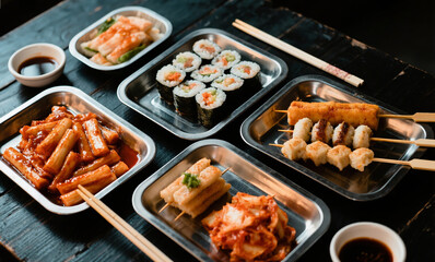 Flatlay of Korean Street Food Spread with Tteokbokki, Kimbap, Fish Cake Skewers, and Kimchi on Metal Trays over a Dark Wood Table