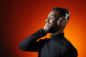Young African man enjoying music with headphones against a vibrant orange background, radiating...