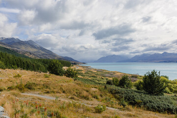 A view of turquoise waters of Lake Pukaki stretching towards hazy mountains under an overcast sky,...