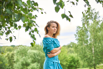 A woman in a blue dress is standing in a grassy field. She is looking at the camera with a serious...