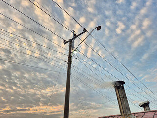 Electric power pole with glowing streetlamp and intersecting wires under cloudy twilight sky with...