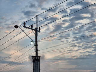 Electric power pole with glowing streetlamp and intersecting wires under cloudy twilight sky with...