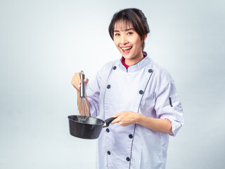 Asian female chef in white uniform holding a black saucepan and metal whisk, smiling...