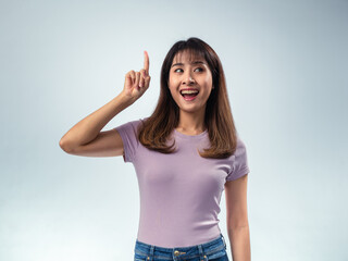 Asian woman in purple t-shirt and jeans smiling brightly while pointing upward with one finger,...