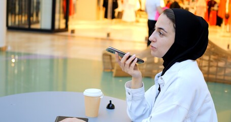 Sitting at a table in a shopping center with a takeaway coffee cup, a young woman wearing a hijab uses her smartphone to record and send voice messages. Muslim with smartphone in hijab. Free Muslim