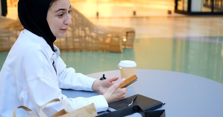 Young muslim woman wearing traditional hijab sitting at a table in a shopping mall, using her smartphone with shopping bags and a coffee next to her. Happy Muslim woman in black hijab
