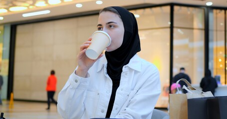 Young muslim woman wearing hijab sitting at table in shopping mall, comfortably browsing smartphone with relaxed smile, embodying modern digital lifestyle and urban connectivity