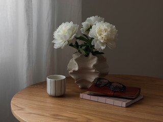 A bouquet of white peonies, a cup of coffee, a notebook on a round wooden table in the living room