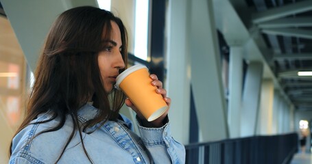 Young woman with long brown hair, wearing a denim shirt, sips coffee from a disposable cup in a modern building with large windows and metal framework