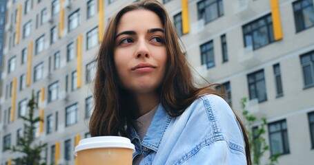Confident young woman with long brown hair, wearing a light blue denim shirt over a beige top, poses and claps her hands in front of a modern residential building with gray and yellow facade