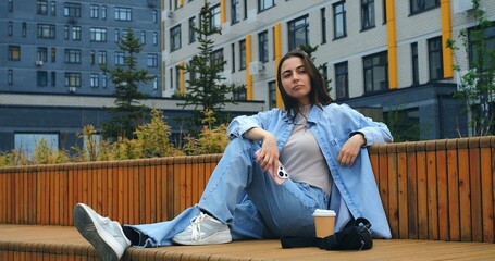 Modern buildings provide the backdrop as a young woman, dressed in casual attire, enjoys a beverage while engaging with her smartphone on a comfortable bench