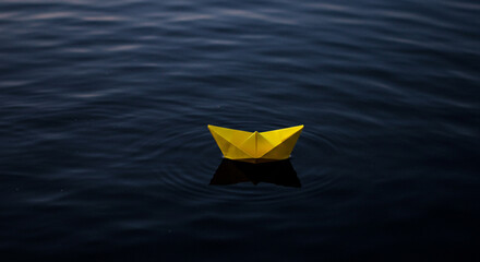 A yellow paper boat floats gently on the water reflecting its shadow in the dark