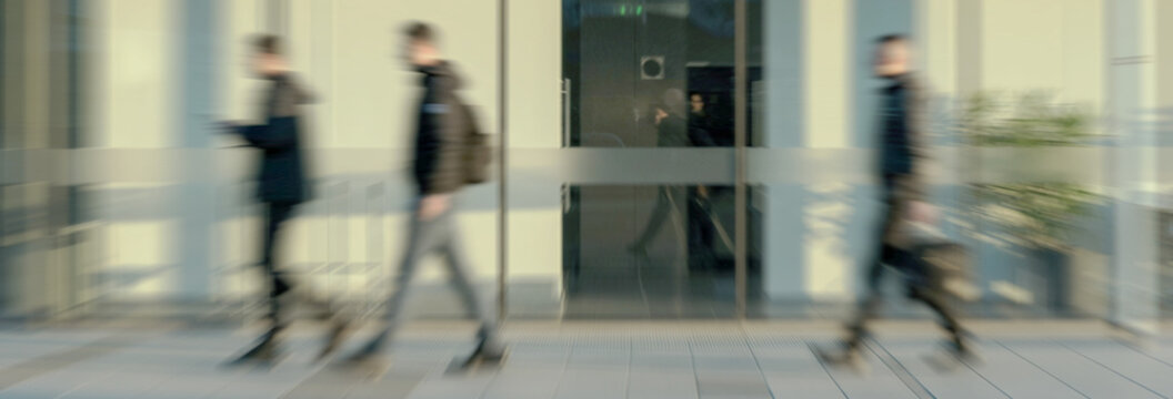 Blurred figures of people are seen walking past glass doors at the entrance of a modern building, conveying a sense of movement and urban activity. - Powered by Adobe