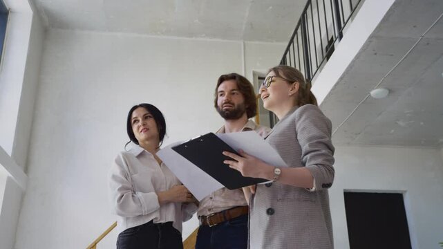 The couple stands with their real estate agent, reviewing architectural plans of a duplex flat in a smooth, low-angle steadicam view