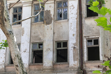 A building with broken windows and cracked walls, surrounded by trees in the city including an old...
