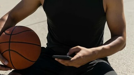 A man sits outdoors holding a basketball and using his phone - Powered by Adobe