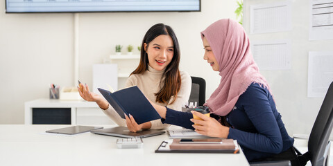 Two asian businesswomen are collaborating on a project, analyzing financial data and taking notes, demonstrating teamwork and expertise in a modern office setting