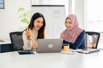 Concentrated young woman in hijab sitting with smiling colleague at table, looking at computer...
