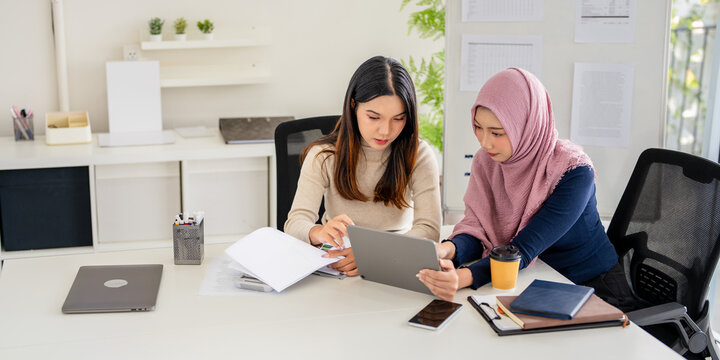 Concentrated young woman in hijab sitting with asian colleague at table, looking at lablet screen, explaining new company job. Focused team leader training female intern.