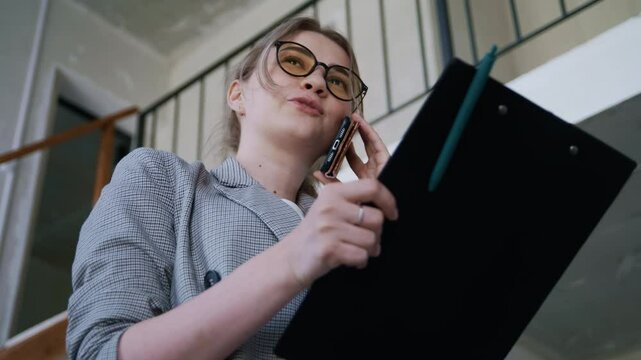 Real estate agent wearing glasses stands in a modern two-level apartment, holding documents and speaking on the phone with a client. Handheld low-angle perspective