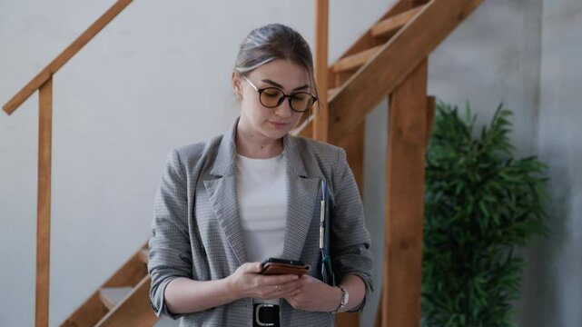 Real estate agent manages business communication while holding paperwork and typing on a smartphone in a bright, unfurnished apartment with visible stairs and a green plant