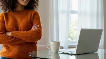 A woman stands confidently with her arms crossed, looking thoughtfully at a laptop on a table