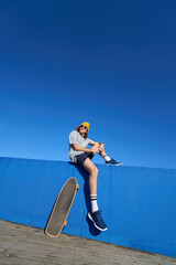 Young skater resting on a blue wall with his skateboard under a clear sky