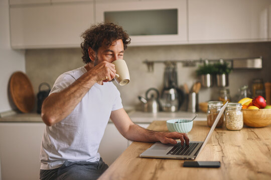 A man sips coffee from a mug while focusing on his laptop in a bright kitchen filled with fruits and breakfast items during the morning.