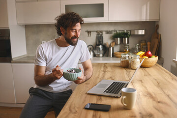 A young man is seated at a wooden kitchen table, eating a bowl of cereal and smiling at his laptop. Fresh fruit and ingredients are visible in the background, indicating a cozy morning.