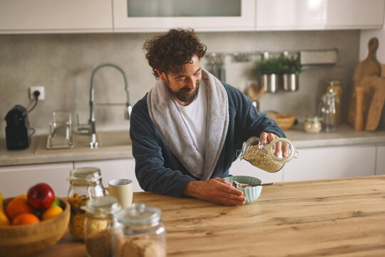 A man in a cozy robe is pouring granola into a bowl in a stylish kitchen. Fresh fruits and jars are placed on a wooden table, hinting at a nutritious breakfast routine.