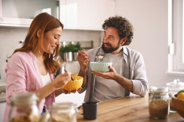 Two people are sharing a joyful breakfast moment in a well-lit kitchen. They are sitting at a wooden table, eating from colorful bowls with smiles on their faces.