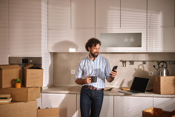 A man stands in a bright kitchen filled with moving boxes, checking his phone and sipping coffee,...
