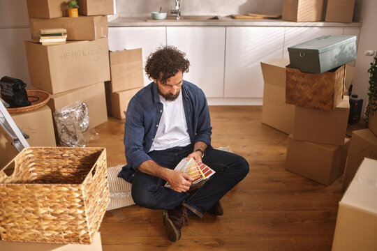 A person sits cross-legged on the floor of a new home surrounded by unpacked boxes, examining different color samples for decorating the interior, clearly in the process of settling in.