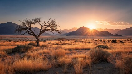 A lone, leafless tree stands silhouetted against a vibrant sunset over a vast, golden-hued desert plain, backed by majestic mountains