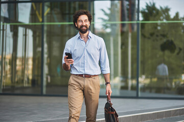 A business professional is smiling as he walks outside a contemporary office building. He is...