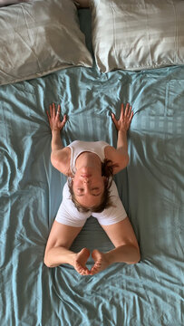 Woman practicing yoga frog pose (Mandukasana) on bed at home, top view. Relaxing morning yoga for flexibility and inner balance.