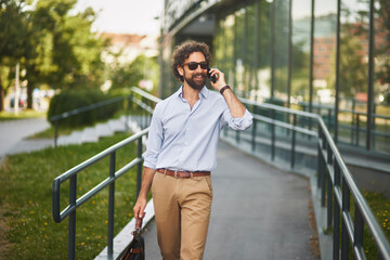 A man in smart casual attire walks confidently along a pathway, engaged in a phone conversation. Sunlight highlights the modern architecture around him.
