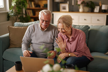 An elderly couple sits on a couch in their living room, smiling and waving at a laptop during a video call. They seem happy and engaged while connecting with family.