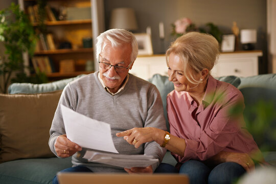 A senior Caucasian couple, sitting on a sofa in their living room, are reviewing documents. The woman points at a paper, while the man holds other papers. They discuss financial matters.