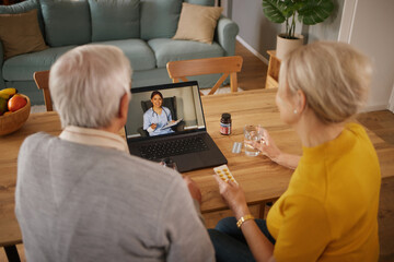 Mature Caucasian couple is sitting at a wooden table, engaging in a telehealth consultation with a doctor who is visible on their laptop screen. They are discussing medication and healthcare options.