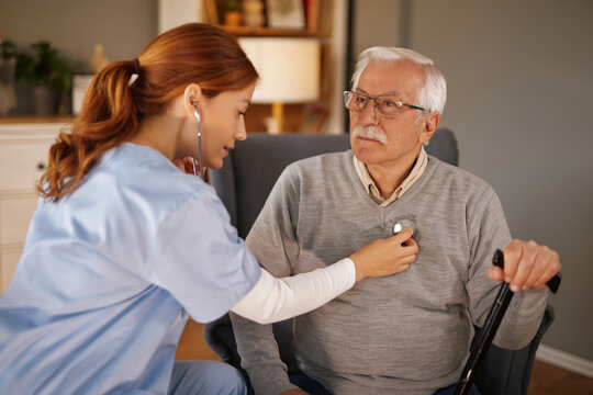 Young female home care nurse uses a stethoscope to examine elderly man with glasses and a walking stick, sitting in an armchair, in his Living Room.
