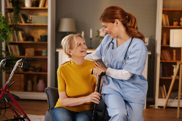 In a cozy living room setting, a home health aide in blue scrubs warmly assists a smiling senior...