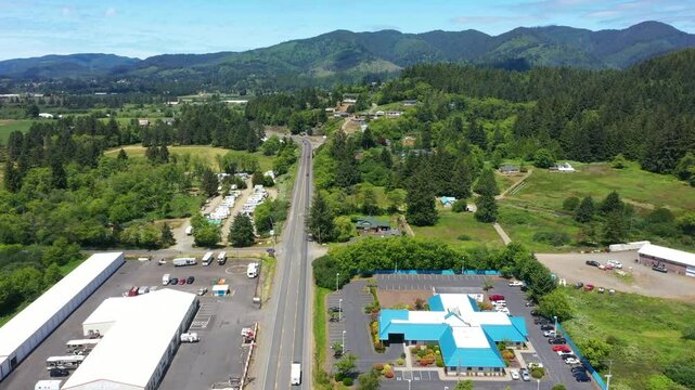 4K aerial drone video of Highway 101 running through Tillamook, Oregon, showing surrounding forested hills, parked vehicles, nearby buildings, and open land on a clear summer day