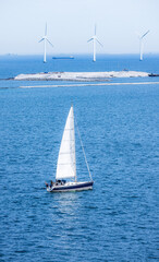 Sailing yacht in the blue waters of Copenhagen with wind turbines in the background on a sunny day, symbolizing clean energy and maritime lifestyle.