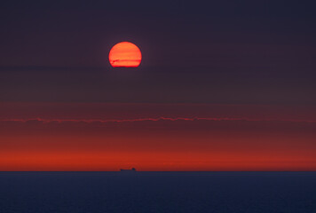 Sunset over the Norwegian Sea with the glowing sun touching the horizon above the silhouette of a distant ship. A dramatic seascape perfect as a bold and warm background image. View from cruise ship