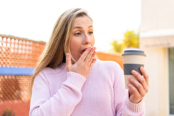 Young blonde woman holding a take away coffee at outdoors with surprise and shocked facial...