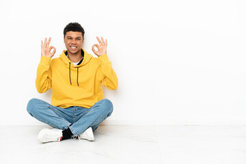 Young African American man sitting on the floor isolated on white background showing an ok sign with fingers