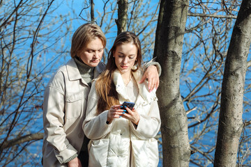 Mature mother and adult daughter are doing selfie by mobile phone in summer park, near lake.